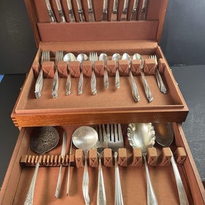 May include: A vintage silverware set in a wooden case. The set includes forks, spoons, knives, and serving utensils. The silverware has a decorative handle design. The case is lined with a brown fabric.