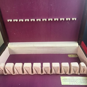 May include: Open wooden silverware storage box with a deep purple interior. The box has slots for utensils and individual compartments. The interior is lined with a soft, light pink fabric. A label is visible inside the box.