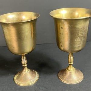 May include: Two matching brass goblets with a polished metallic finish. Each vessel features a wide, flared rim, a smooth rounded bowl, a slender stem with a central decorative ring detail, and a circular base for stability on flat surfaces.