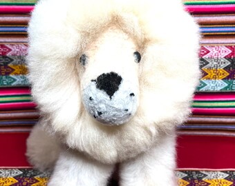 Op de afbeelding: Wit, pluche alpaca leeuwenknuffel die op een kleurrijke geweven stoffen achtergrond zit.