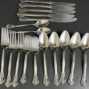 Könnte beinhalten: Eine Sammlung von Vintage-Besteck, darunter Gabeln, Löffel und Messer, auf einer dunklen Oberfläche angeordnet. Das Besteck hat verzierte florale Details an den Griffen. Die Löffel, Gabeln und Messer sind aus glänzendem, silberfarbenem Metall gefertigt.