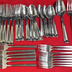 May include: A collection of stainless steel silverware, including forks, spoons, and knives, arranged on a red surface. The silverware has a polished finish, reflecting the light. The forks have four tines, and the spoons have a rounded bowl.