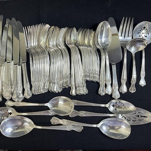 May include: A large collection of silver-toned flatware including dinner knives, forks, spoons, and serving utensils. The set features ornate decorative handles with intricate detailing and includes specialized pieces like a slotted spoon and cake server.