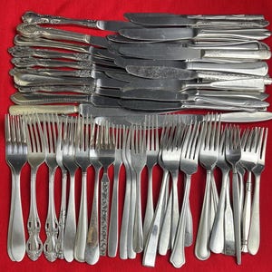 Puede incluir: Una colección de cubiertos antiguos, que incluye tenedores y cuchillos, dispuestos sobre una superficie roja. La cubertería presenta diversos diseños de mangos y estilos de hojas, mostrando una gama de patrones y texturas. Ideal para coleccionistas de antigüedades.