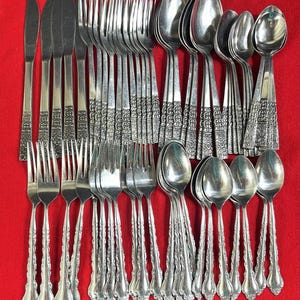 May include: A collection of vintage silverware, including knives, forks, and spoons, arranged on a red surface. The silverware has ornate, detailed handles and a shiny, reflective surface, suggesting a formal dining set.