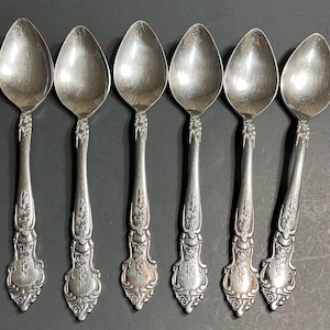 Puede incluir: Seis cucharaditas de plata con mangos ornamentados. Las cucharas tienen un acabado pulido y están dispuestas en fila.