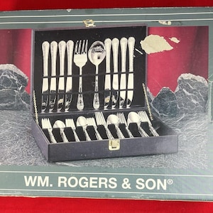 May include: A silver-coloured cutlery set presented in a black case with a gold clasp. The set includes forks, spoons, and knives. The box is labelled "WM. ROGERS & SON®" in white text on a dark green background.