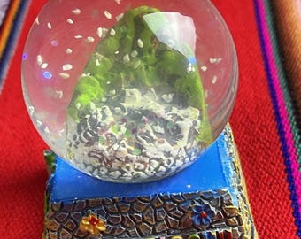 May include: A snow globe featuring a green mountain scene inside a clear glass sphere. The base is blue and brown, with the words "MACHUPICCHU CUSCO PERU" in gold lettering. Colorful flowers decorate the base.