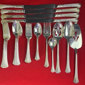 Puede incluir: Una colección de cubiertos antiguos, que incluye cuchillos, tenedores, cucharas y utensilios para servir. La cubertería plateada está dispuesta sobre un fondo rojo. El juego incluye varios tamaños de cucharas, tenedores y una espátula para tartas.