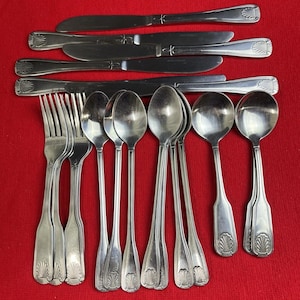 May include: A collection of stainless steel silverware, including knives, forks, and spoons, arranged on a red surface. The silverware has a decorative shell design on the handles.