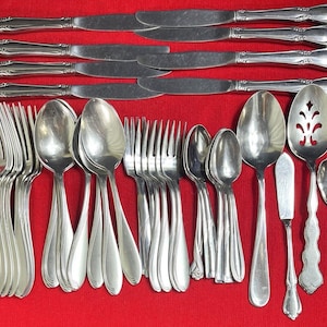 Può includere: Una collezione di posate color argento, tra cui coltelli, forchette, cucchiai e utensili da portata, disposte su una superficie rossa. I coltelli hanno un design decorativo sul manico. Le forchette, i cucchiai e i pezzi da portata variano per dimensioni e forma.