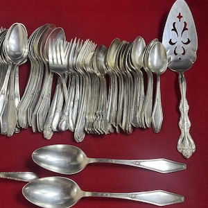 Könnte beinhalten: Eine Sammlung von Vintage-Silberbesteck, darunter Löffel, Gabeln und ein Tortenheber, angeordnet auf einer roten Oberfläche. Das Besteck hat eine glänzende, silberne Oberfläche mit aufwendigen Griffdetails. Der Tortenheber hat ein dekoratives, durchbrochenes Design.