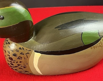 May include: A hand-painted wooden duck figurine. The duck features a brown head with a green eye patch, a tan beak, and a body painted in shades of green, brown, and tan. The duck is resting on a red surface.