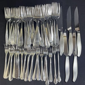 Può includere: Una collezione di posate in metallo color argento composta da numerose forchette di varie dimensioni e design, insieme a quattro coltelli da tavola con manici lisci e semplici. Gli utensili sono disposti in gruppo e presentano finiture metalliche.