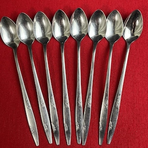 May include: Eight silver spoons displayed on a red surface. Each spoon features a long handle with a floral pattern near the bowl. The spoons are reflective, showing the surrounding environment.