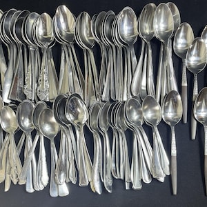 Può includere: Una vasta collezione di cucchiai in metallo color argento assortiti con vari design del manico, inclusi stili lisci, goffrati e bicolore con accenti scuri. I cucchiai sono disposti in un gruppo denso e sovrapposto.