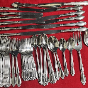 Pode incluir: Um conjunto completo de talheres prateados, incluindo facas, garfos, colheres e utensílios para servir. A prataria está disposta sobre uma superfície vermelha. As alças têm um design decorativo e ornamentado.