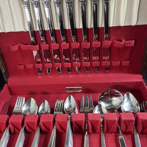 May include: A set of silver-colored flatware, including knives, forks, and spoons, neatly arranged in a red velvet-lined wooden case. The handles feature an ornate floral design. The case is open, revealing the silverware's arrangement.