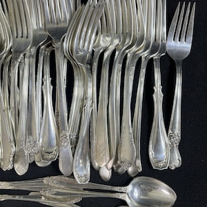 May include: A collection of silver-toned metal forks and spoons arranged in a group. The forks feature various decorative embossed patterns on the handles, while the spoons have smooth, oval-shaped bowls and matching ornate handle designs.