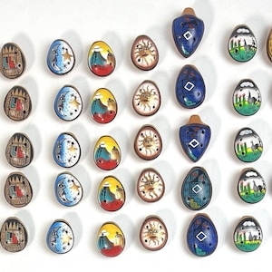 Puede incluir: Una colección de ocarinas de cerámica pintadas a mano en varios colores y diseños. Las ocarinas presentan patrones de paisajes, celestes y geométricos. Algunas tienen forma de lágrima, otras son ovaladas. La paleta de colores incluye azul, amarillo, marrón y negro.