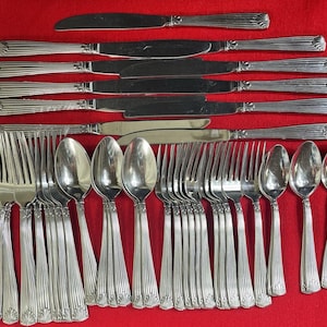 May include: A complete set of silver-colored flatware, including knives, forks, and spoons, arranged on a red surface. The utensils have a decorative, ridged design on the handles. The knives have a smooth, polished blade.