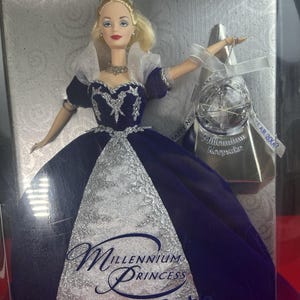 Peut inclure: Poupée Barbie, la Princesse du Millénaire, dans une boîte d'exposition. La poupée porte une robe bleu foncé avec des accents argentés et un diadème. La boîte comprend un souvenir argenté avec un globe et le texte "Happy New Year 2000."