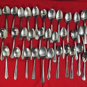 Könnte beinhalten: Eine Ansammlung verschiedener silberner Löffel, die auf einer roten Oberfläche angeordnet sind. Die Löffel haben unterschiedliche Griffdesigns und Schalenformen, die eine Vielzahl von Stilen und Größen zeigen. Die Löffel bestehen aus glänzendem Metall.