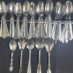 May include: A collection of various silver-toned metal spoons arranged in two rows. The top row features multiple spoons with ornate handles and smooth bowls, while the bottom row displays five distinct spoons with varied handle designs and shapes.