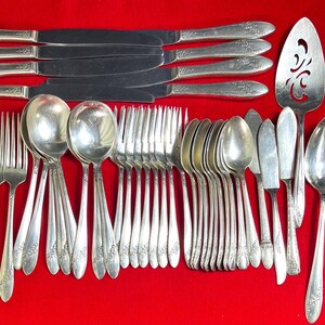 May include: A set of vintage silverware, including knives, forks, spoons, and serving utensils, arranged on a red surface. The silverware has a floral design on the handles. Includes a pie server.