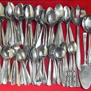 May include: A collection of various silver spoons, serving spoons, and a cake server, all with different handle designs. The spoons are arranged on a red surface, showcasing their polished surfaces and diverse styles. The assortment includes both everyday and decorative pieces.