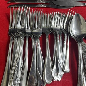 May include: A collection of stainless steel silverware, including forks, knives, and spoons, arranged on a red surface. The silverware has decorative handles and a shiny finish, suitable for formal dining.