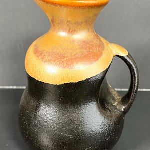 May include: A brown and black ceramic pitcher with a wide mouth and a handle. The pitcher has a textured surface and a glossy glaze.