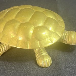 May include: A gold-colored brass turtle figurine featuring a detailed, segmented shell pattern, a smooth head with defined eyes, and four legs with visible claw markings. The object has a polished metallic finish and a realistic, rounded shape.