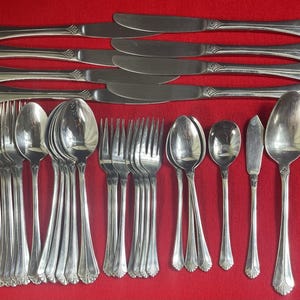 May include: A collection of stainless steel cutlery on a red surface. The set includes knives, forks, spoons, and butter knives. The cutlery features a decorative handle design.