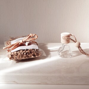 May include: A small, clear glass bottle with a heart-shaped design, hanging from a brown twine cord. The bottle has a wooden cap with a natural finish. The bottle is packaged in a clear plastic bag with brown shredded paper and a small brown tag that says "Candles".