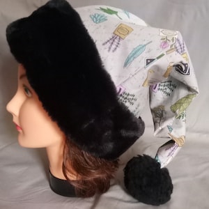 May include: A black faux fur hat with a long, pointed crown. The crown is made of white fabric with a floral print featuring herbs, leaves, and stars. The hat has a black pom-pom on the end of the crown.