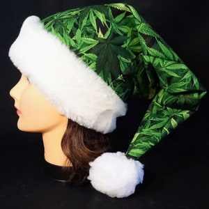 May include: A green and white Santa hat with a marijuana leaf print. The hat has a white fur trim and a white pom-pom.