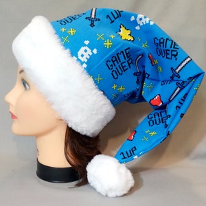 May include: A blue Santa hat with a white faux fur trim and a white pom-pom. The hat is patterned with pixelated video game graphics, including swords, skulls, and the words "GAME OVER" and "1UP".
