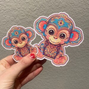 May include: Two colorful monkey stickers with intricate patterns and glitter accents. The larger sticker is pink and blue, while the smaller sticker is pink and orange.