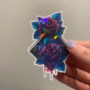 May include: A holographic sticker featuring two purple roses with blue-green leaves. The roses are arranged in a diamond shape with a black background and dripping paint.