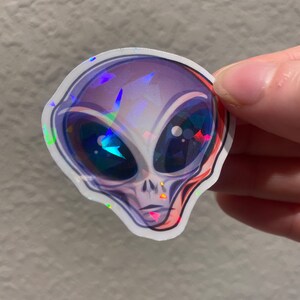 May include: A holographic sticker of a grey alien with large eyes and a serious expression.