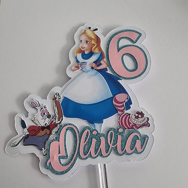 Peut inclure: Décor de gâteau sur le thème d'Alice au pays des merveilles avec un numéro 6 rose, un lapin blanc, le chat du Cheshire et le texte "Olivia" en paillettes roses et turquoise.
