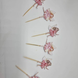May include: Six pink and white fairy cupcake toppers with brown hair and wings. The fairies are on wooden skewers.