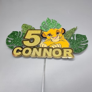 May include: Gold glitter cake topper featuring a cartoon lion cub, the number 5, and the name "CONNOR" surrounded by green glitter leaves.