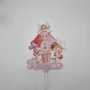 May include: A pink and white fairy cake topper with a mushroom house, two fairies, and the number 6. The name "Margaux" is written in silver glitter on the bottom.