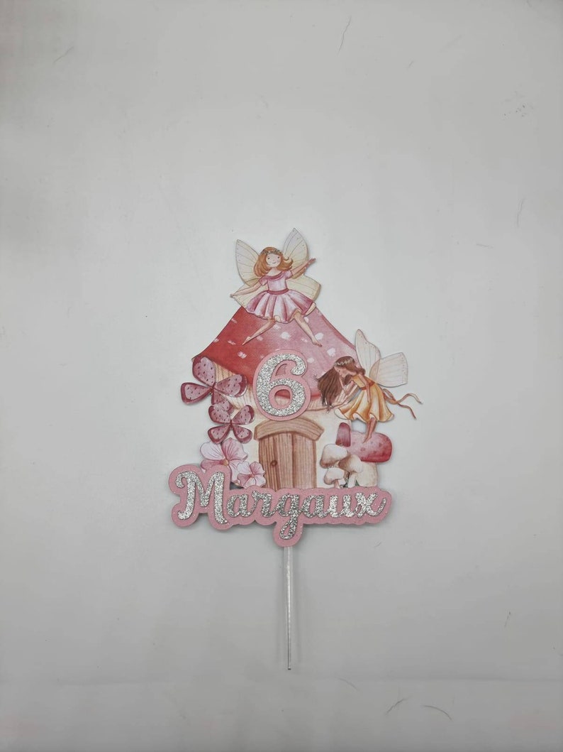 May include: A pink and white fairy cake topper with a mushroom house, two fairies, and the number 6. The name "Margaux" is written in silver glitter on the bottom.