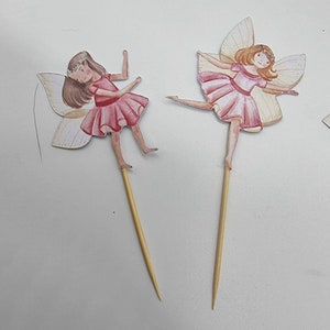 May include: Two fairy cake toppers with pink dresses and wings. The fairies are holding a stick in their hands. The toppers are made of paper and are attached to wooden sticks.