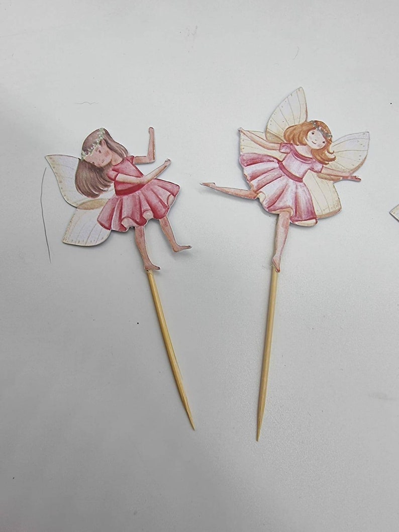 May include: Two fairy cake toppers with pink dresses and wings. The fairies are holding a stick in their hands. The toppers are made of paper and are attached to wooden sticks.