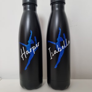 May include: Two black stainless steel water bottles with a blue glitter dancer silhouette and personalized names "Harper" and "Isabelle" in white lettering.