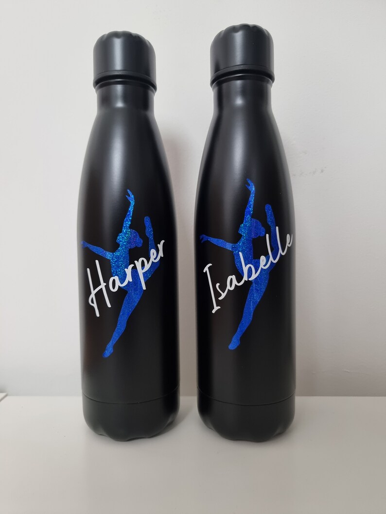 May include: Two black stainless steel water bottles with a blue glitter dancer silhouette and personalized names "Harper" and "Isabelle" in white lettering.